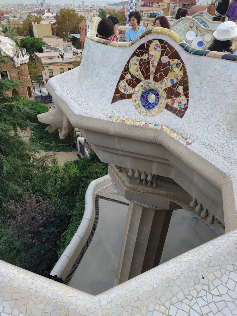 [Alt text: A group of tourists sits and talks on an elaborate balcony, surrounded by a low wall of multicolored tiles. The balcony is supported by pillars which go far down into a green area full of trees. Further in the distance, various different city buildings are visible, and in the far distance there is a faint glimpse of the sea.]
